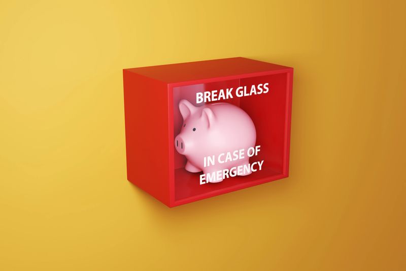 Pink piggy bank in a red box with a glass printed with the instruction BREAK GLASS IN CASE OF EMERGENCY on a yellow wall. Illustration of the concept of emergency fund and financial buffer