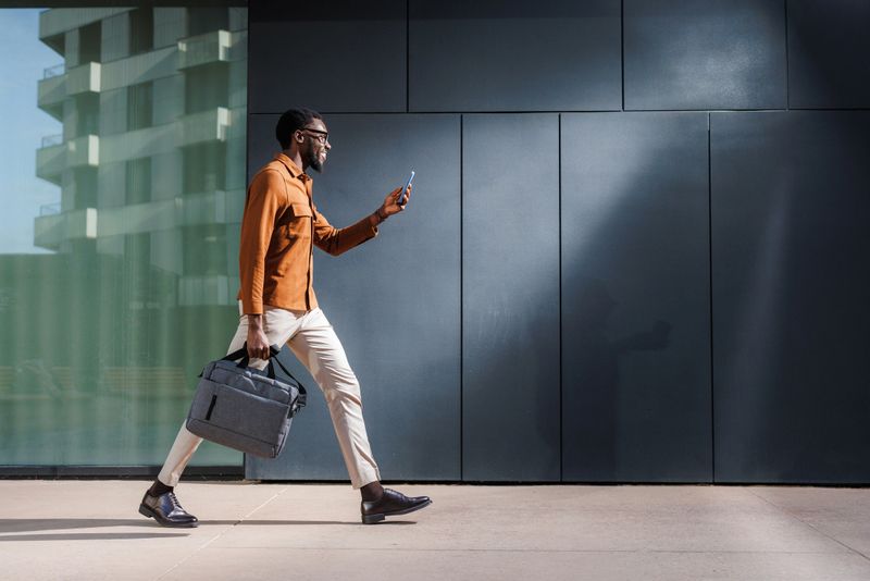 A focused man walks confidently outside a modern office building while checking his smartphone. He carries a stylish briefcase, showcasing productivity and urban working life.