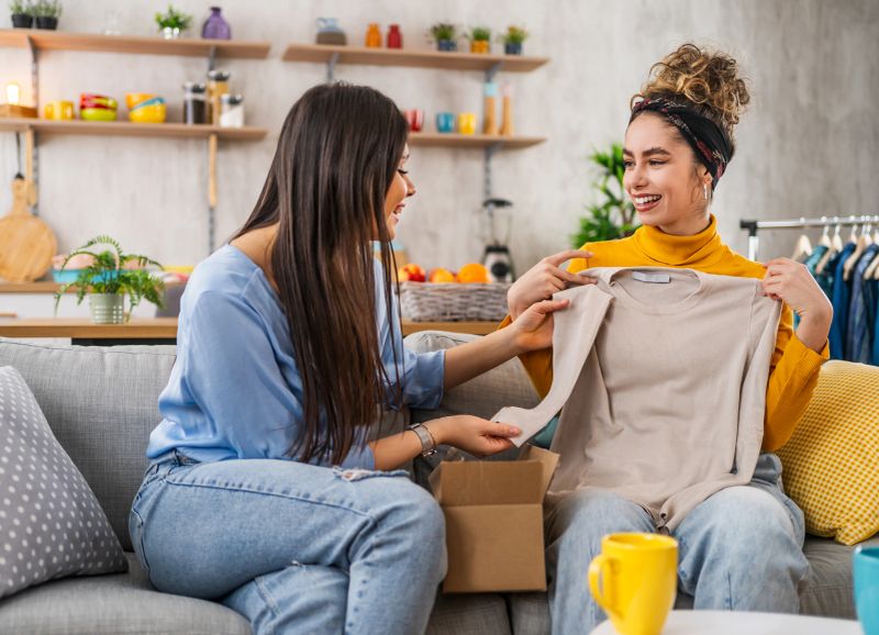 Two female friends are unpacking clothes from a delivery box, checking the quality of the product at home