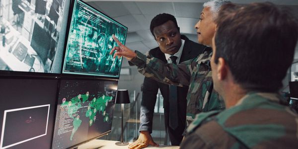Military personnel and a man in a suit analyzing global data on multiple screens.