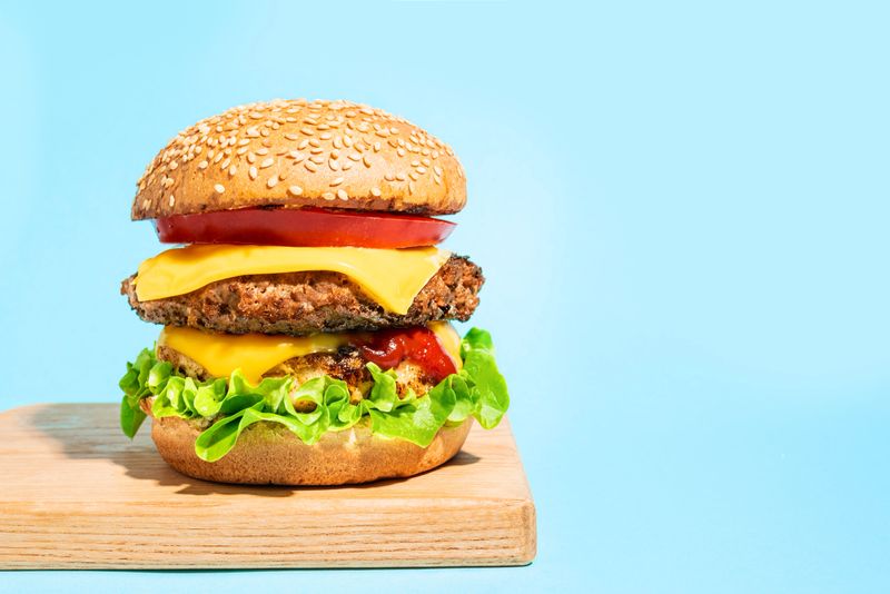 Double cheeseburger with lettuce, tomato, ketchup and melted american cheese on blue background, copy space. Big burger with extra meat. Hamburger party design.