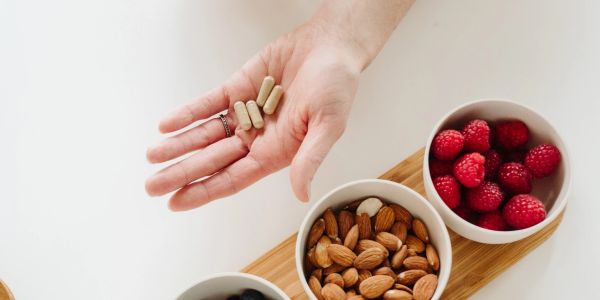 Hand holding capsules with bowls of nuts and berries nearby.