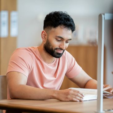 Man in a pink shirt reading a book at a table.