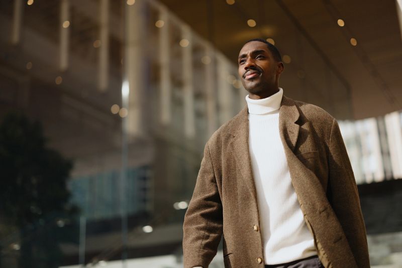 A confident man in trendy attire walks through a modern building exterior in soft lighting, reflecting sophistication and urban energy.