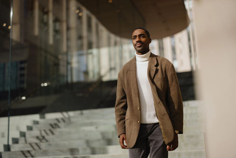 Confident man in stylish attire walking outdoors in a city setting with modern architecture, showcasing elegance and sophistication in a professional urban backdrop.