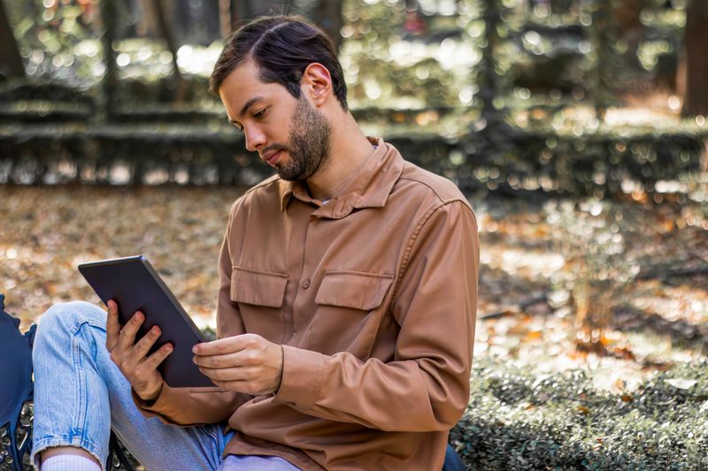 Young man sitting on a park bench, reading an e book on a tablet while leaning forward, highlighting bad posture and potential neck discomfort amid the serene autumn surroundings