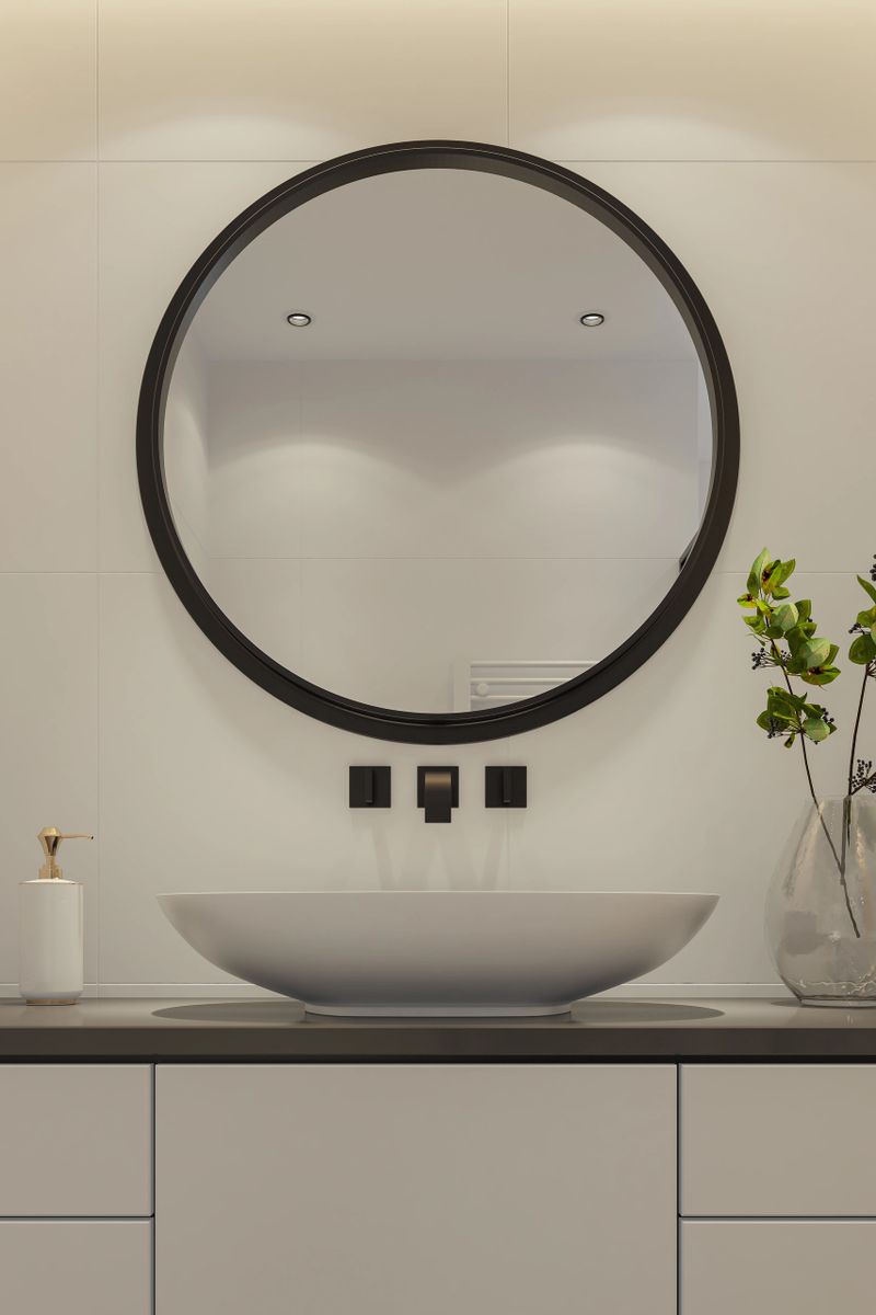 Stylish and minimalistic bathroom interior featuring a wall-mounted oval sink with a sleek black faucet, set against matte white large-format tiles. A floating cabinet adds functionality without compromising the clean aesthetic, while a large round mirror enhances the sense of space.Perfect visual for interior design inspiration, bathroom remodeling ideas, tile and fixture brand promotion, or showcasing modern bathroom accessories and trends.