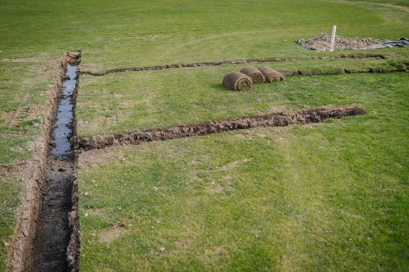 A trench is dug in a grassy field with rolled turf laid nearby, likely for irrigation or drainage installation during landscaping work.