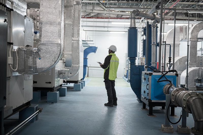 professional Asian engineer or technician in safety gear performs a routine inspection using a digital tablet inside a modern industrial facility. Surrounded by high-tech piping and equipment, he checks operational systems in a controlled, clean, and safe work environment.