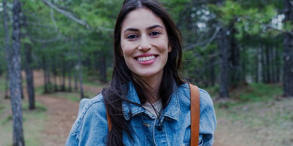 Woman with brown hair and eyes smiling at the camera, wearing jean jacket & a backpack in the forest