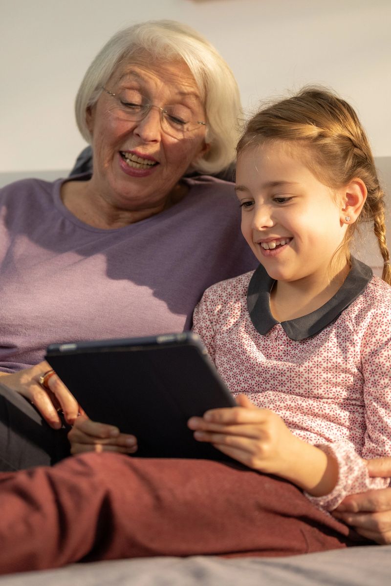 Close-up portrait showing elderly Caucasian woman and young girl with braided hair sharing genuine laughter while viewing tablet device together on comfortable sofa in warm-lit interior.