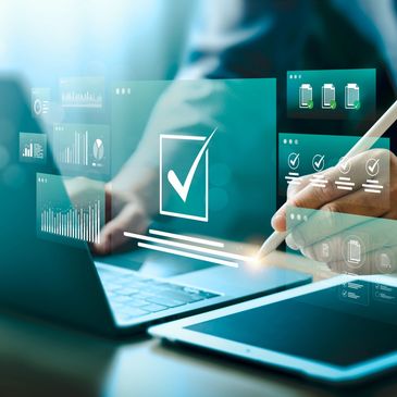 Hand using a stylus on a tablet with digital checklists and graphs hovering over a laptop.