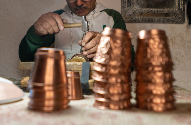 Skilled artisan engraving decorative designs onto a copper cup using a hammer and chisel. The focus is on the craftsman’s precise hands and the detailed metalwork, with stacks of finished copper cups in the foreground.
