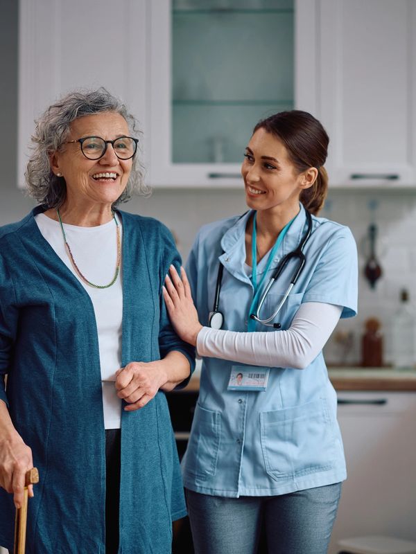A nurse assists a smiling elderly woman using a cane in a bright kitchen.