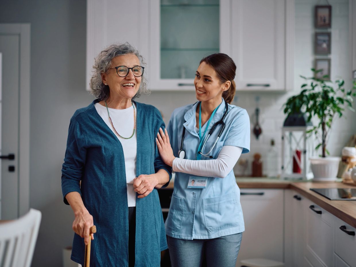Nurse assisting elderly woman with a cane in the kitchen.