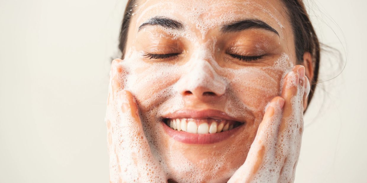 Woman washing face with foamy cleanser, eyes closed, smiling.