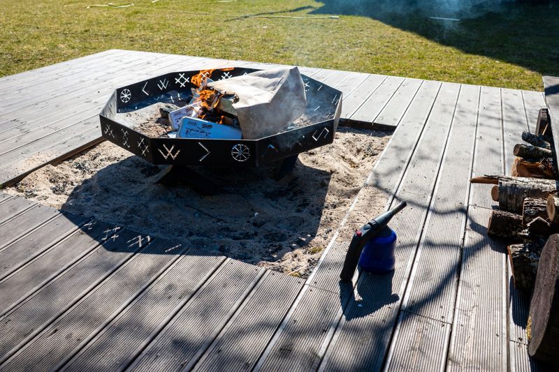 A fire pit with geometric designs burns on a wooden deck surrounded by sand. A blue torch lighter, firewood, and grassy area are visible under sunlight.