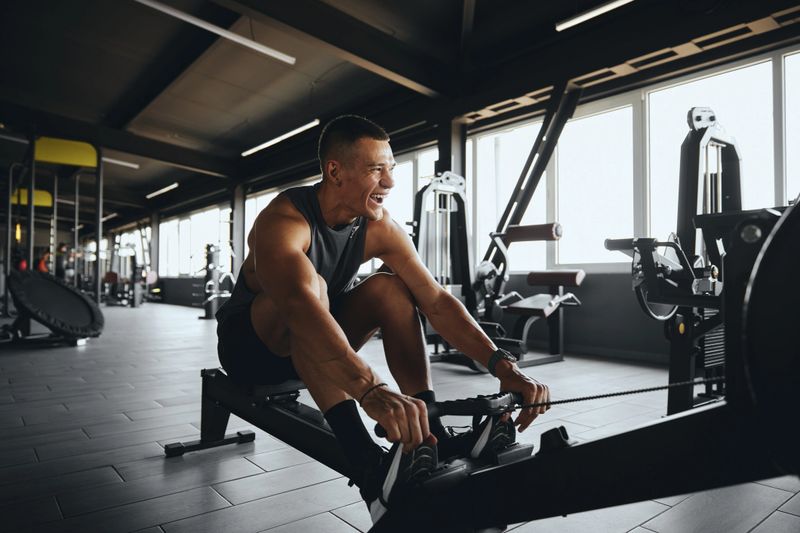 Cheerful attractive man smiling while using rowing machine in contemporary gym. Fit young man working out on rowing equipment at fitness center. Weight loss and cardio exercise concept