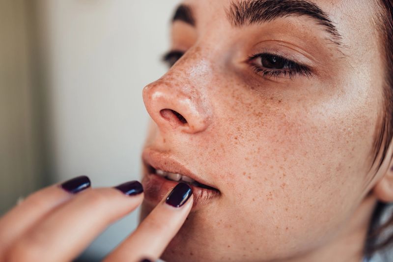 Close-up of a make-up artist's hand applying dark lipstick on a freckled model's lips during a make-up session