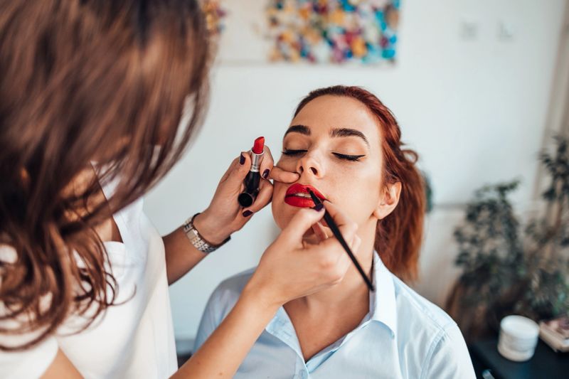 Professional makeup artist applying red lipstick on a client's lips with a brush in a beauty salon