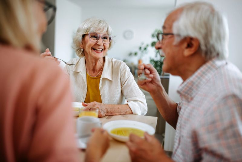 A senior couple shares a peaceful lunch with their grown daughter in their home. The family sits together at the table, enjoying a meal and quality time