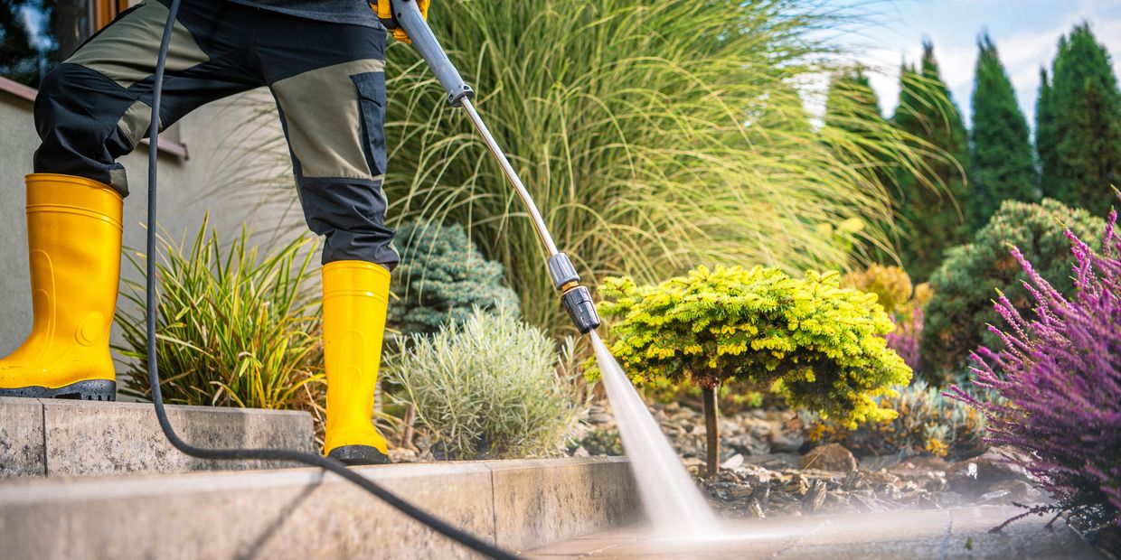 Person in yellow boots power washing an outdoor stone surface near plants.