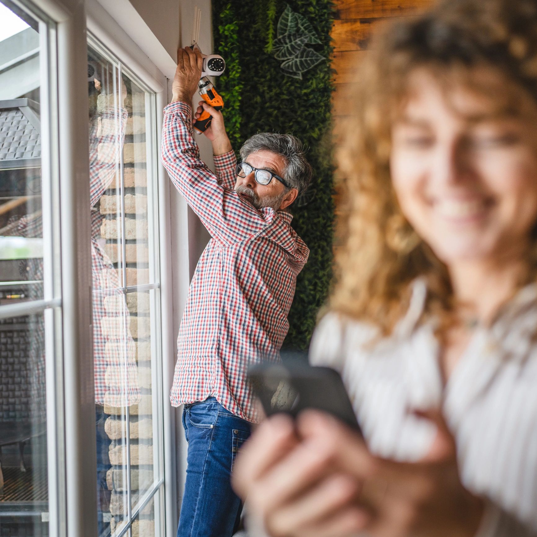 Man installs security camera indoors while woman smiles using phone.