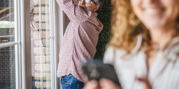Man installs security camera indoors while woman smiles using phone.