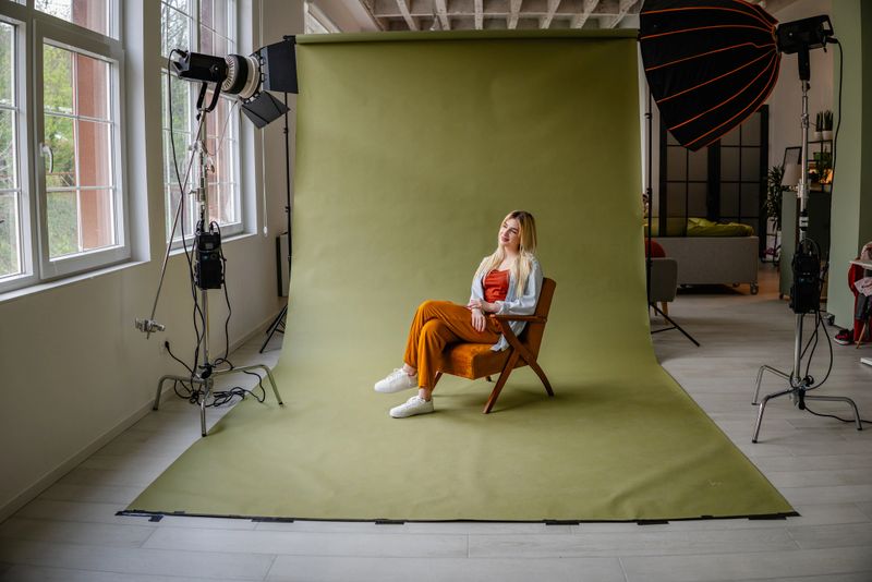 A young blonde woman sits on a wooden chair in a stylish studio setting, showcasing a modern outfit. The cheerful atmosphere is enhanced by natural light from large windows.