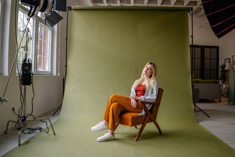 A young blonde woman sitting gracefully in a stylish chair, radiating confidence and joy in a modern studio environment with soft natural lighting and a vibrant backdrop.