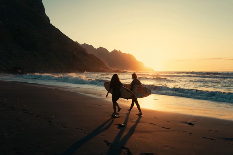Two surfers walk toward the ocean at sunset on a black sand beach in Taganana, Tenerife, with dramatic cliffs and waves in the background.