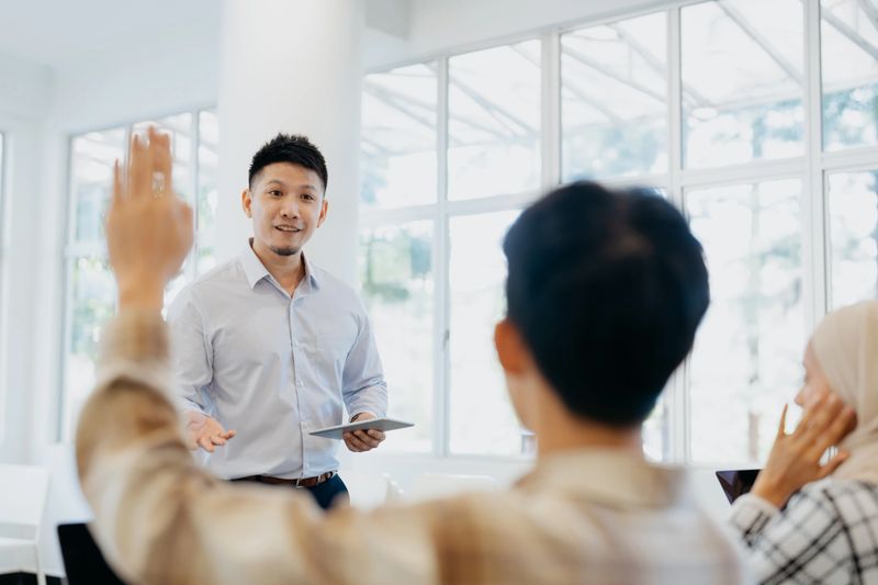 Asian lecturer stands confidently at the front of the room, holding a tablet, while a student in the foreground enthusiastically raises their hand to participate.