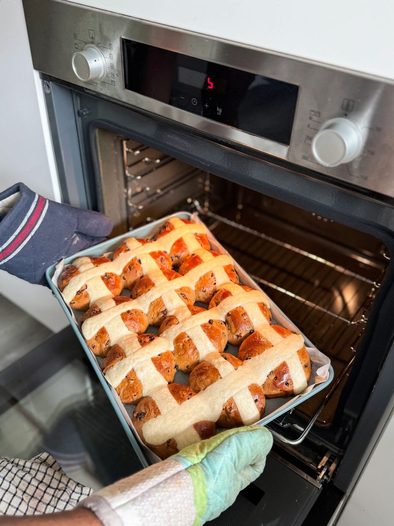 Stock photo showing close-up view of rows of freshly baked, homemade, Easter hot cross buns, being removed from a hot oven shelf, in greaseproof paper lined baking tray. These traditional spiced sweet buns are made and sold over the Easter period, with the cross symbol on the top being made from a flour and water paste, and symbolic to Christianity. Home baking concept.