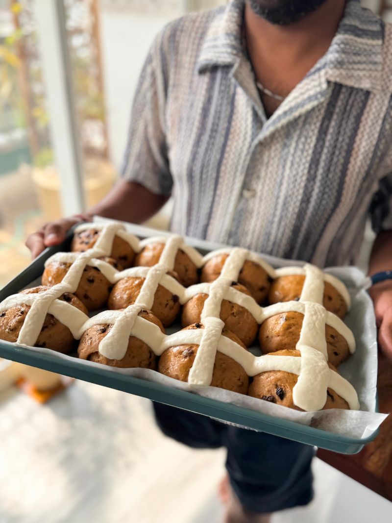 Stock photo showing close-up view of an unrecognisable man carrying a greaseproof paper lined baking tray containing rows of homemade, Easter hot cross buns. These traditional spiced sweet buns are made and sold over the Easter period, with the cross symbol on the top being made from a flour and water paste, and symbolic to Christianity. Home baking concept.