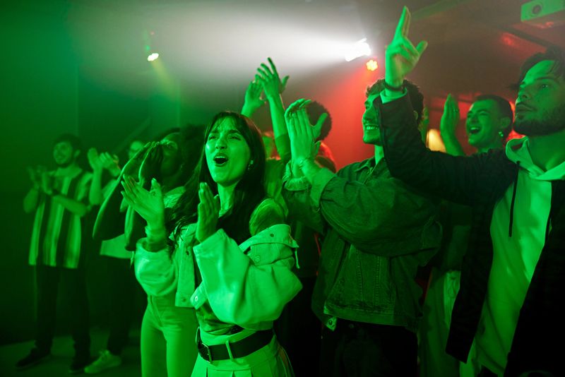 Casually dressed music lovers in their 20s standing side by side on dance floor and showing their appreciation for off-camera jazz band.