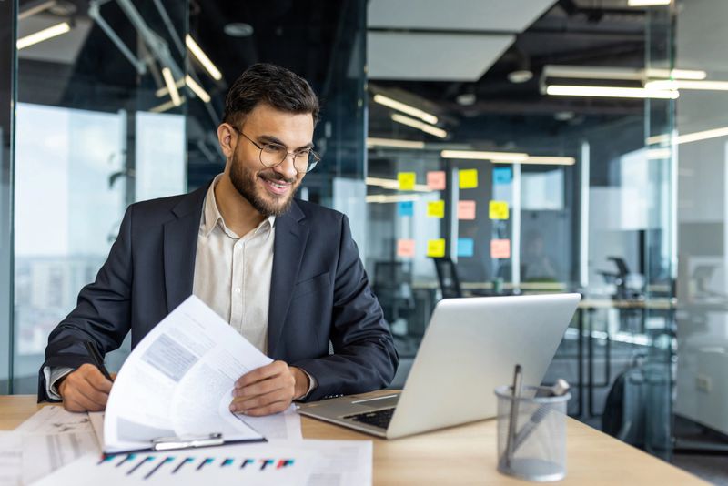 A smiling businessman reviews documents, with a laptop open on his desk in a modern office setting, ready to work.
