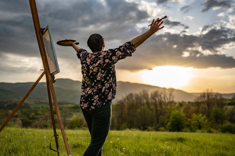 A person in a floral shirt paints outdoors in a countryside setting at sunrise, enjoying the serene beauty of nature.