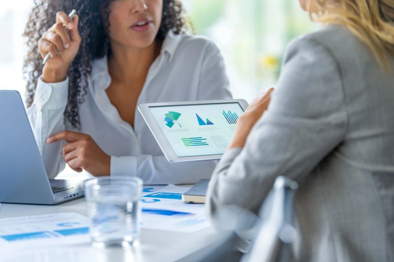 Two business women meeting and looking at financial data, charts and graphs on a digital tablet and a laptop computer. There is paperwork on the table showing finance charts and graphs