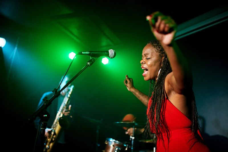 Side view of mature Black musician in red dress standing at microphone, arms raised, drummer and saxophonist in background.