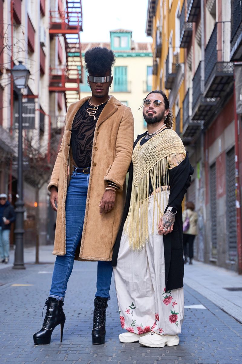 Two non binary people wearing modern fashion outfits are posing together on a city street