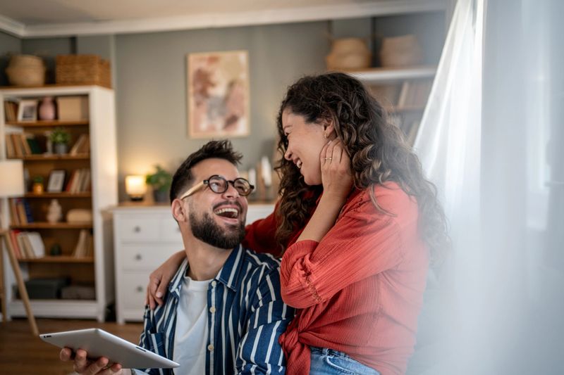 Young couple enjoying time together at home, laughing and using a digital tablet. Embracing connection and comfort in their cozy living room, filled with natural light