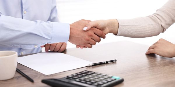 Two people shaking hands over a desk with paperwork and a calculator.