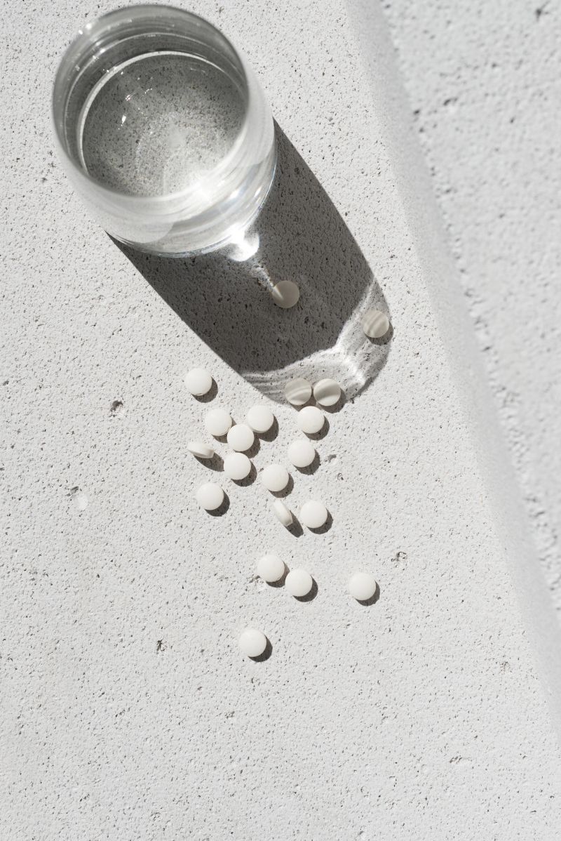 White vitamin pills and glass with pure water on neutral concrete background with aesthetic sunlight shadow. Minimal beauty, wellness or pharmacy concept. Purity and simplicity scene, top view
