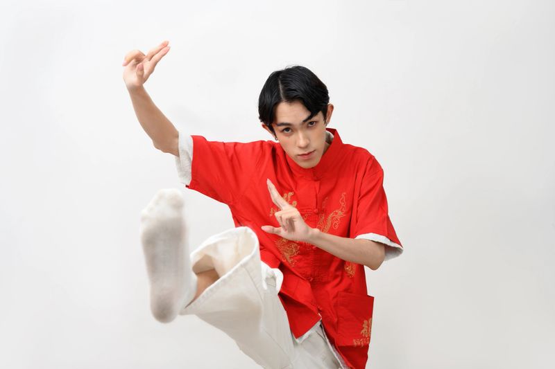 A young Asian teenage boy dressed in a traditional red Chinese outfit performs a high kick and focused hand gestures, embodying the power of kung fu in a studio over a white background.