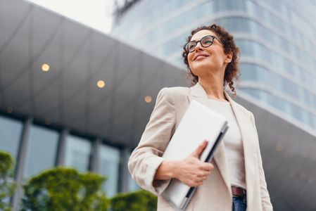 Confident young woman holding a laptop outside a modern office building.