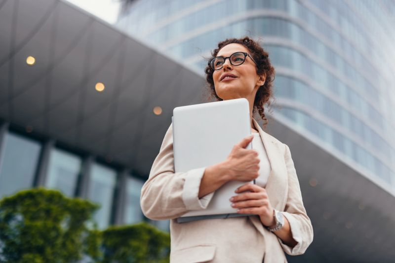 Smiling businesswoman in her 40s is holding a laptop in her arms while looking away and thinking about future projects in front of an office building