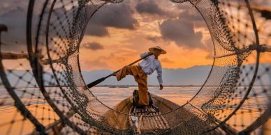 Fisherman balancing on a boat framed by a fishing net at sunset.