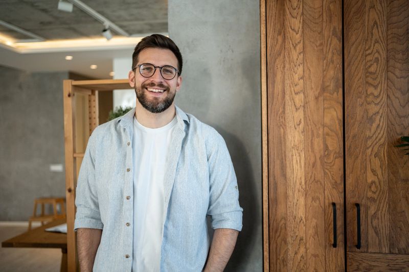 Portrait of a young entrepreneur standing confidently in a modern office. Smiling and looking at the camera, dressed in casual attire and stylish eyeglasses, radiating positivity and ambition