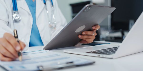 Doctor in blue scrubs holding a tablet, with a laptop on the desk.