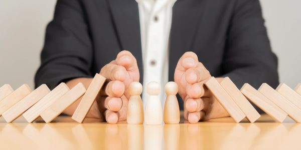 Hands protect wooden figurines from falling dominoes.